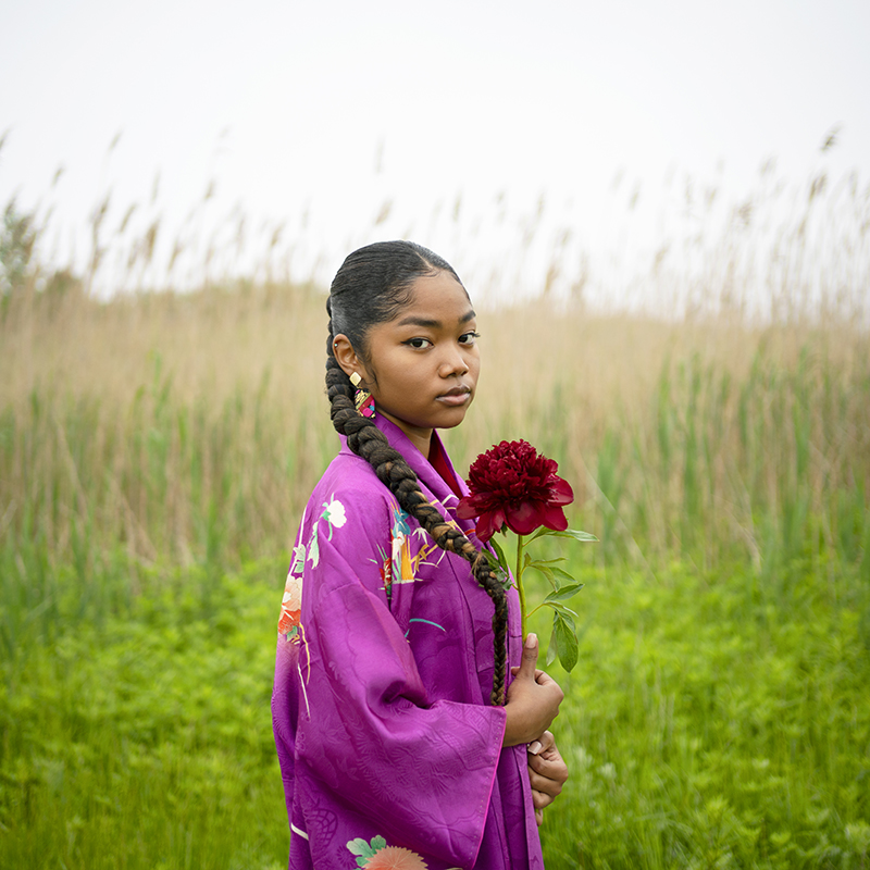 Hidemi Takagi, Mikua from the "IDENTITIES" series, 2022, Digital C Print on the textile. Shows a colored girl wearing purple. She has large, colorful earrings and is holding a red flower.