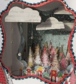 This is a wintery diorama. It is made out of a box that has been cut open in the middle. Inside, there are patterned trees, a line of houses, and hanging clouds.
