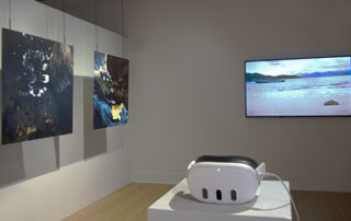 An installation view of ETA at VisArts. There is a VR headset in the foreground and a TV screen displaying a beach in the back. On the left, there are two abstract paintings hanging from the ceiling. Shown in courtesy of VisArts.