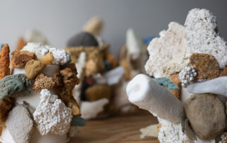 Close-up of an abstract sculptural arrangement made from assorted natural and artificial materials attached to white paper cups. The objects include porous rocks, foam, sponge-like fragments, Styrofoam, and earthy-toned stones in shades of brown, white, gray, and muted turquoise. The textures range from rough and jagged to smooth and rounded, creating a clustered, coral-like appearance on a wooden surface with other similar sculptures blurred in the background.