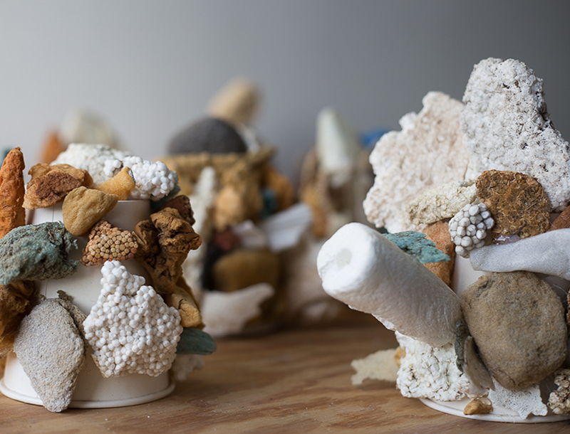 Close-up of an abstract sculptural arrangement made from assorted natural and artificial materials attached to white paper cups. The objects include porous rocks, foam, sponge-like fragments, Styrofoam, and earthy-toned stones in shades of brown, white, gray, and muted turquoise. The textures range from rough and jagged to smooth and rounded, creating a clustered, coral-like appearance on a wooden surface with other similar sculptures blurred in the background.