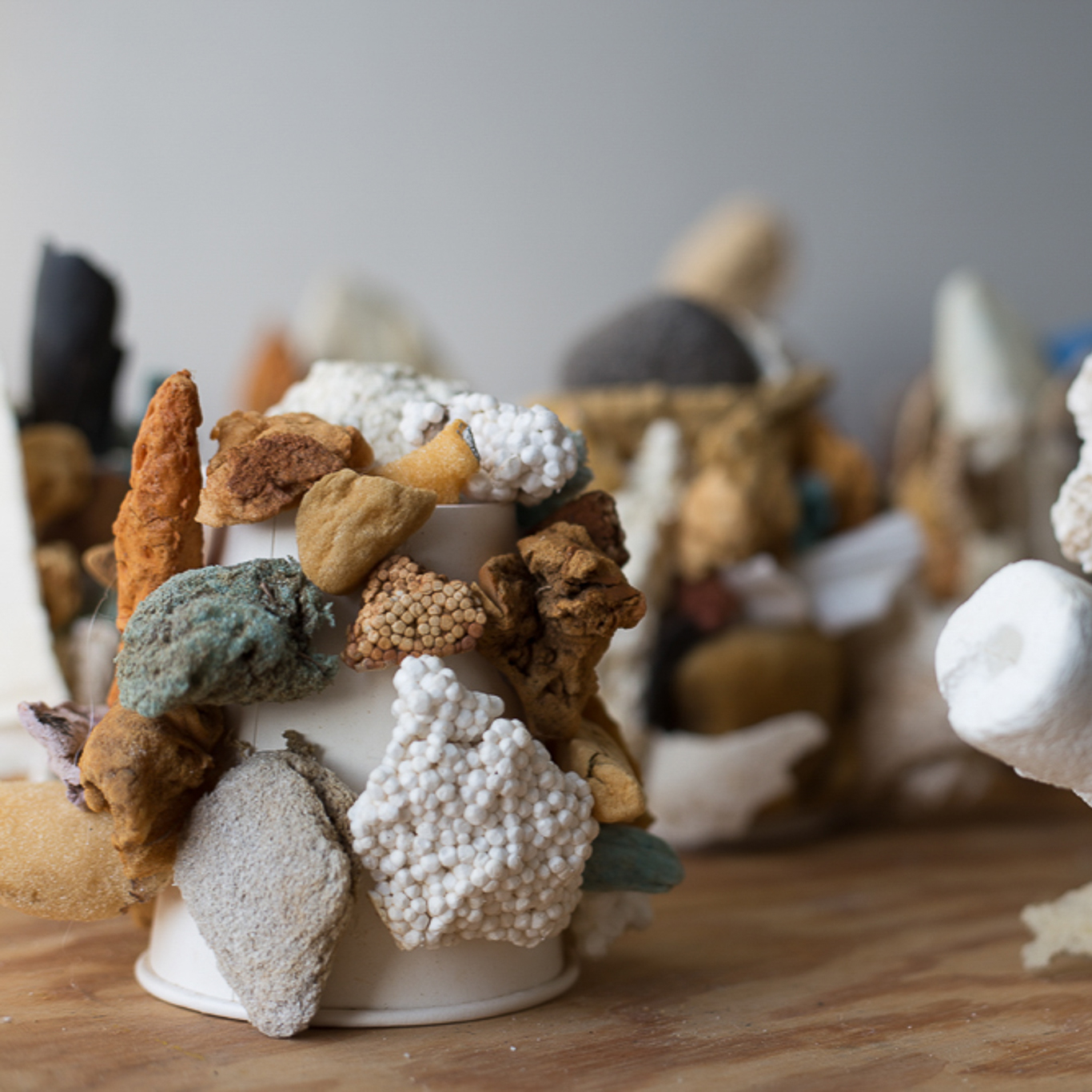 03_conglomeratesq Close-up of an abstract sculptural arrangement made from assorted natural and artificial materials attached to white paper cups. The objects include porous rocks, foam, sponge-like fragments, Styrofoam, and earthy-toned stones in shades of brown, white, gray, and muted turquoise. The textures range from rough and jagged to smooth and rounded, creating a clustered, coral-like appearance on a wooden surface with other similar sculptures blurred in the background.
