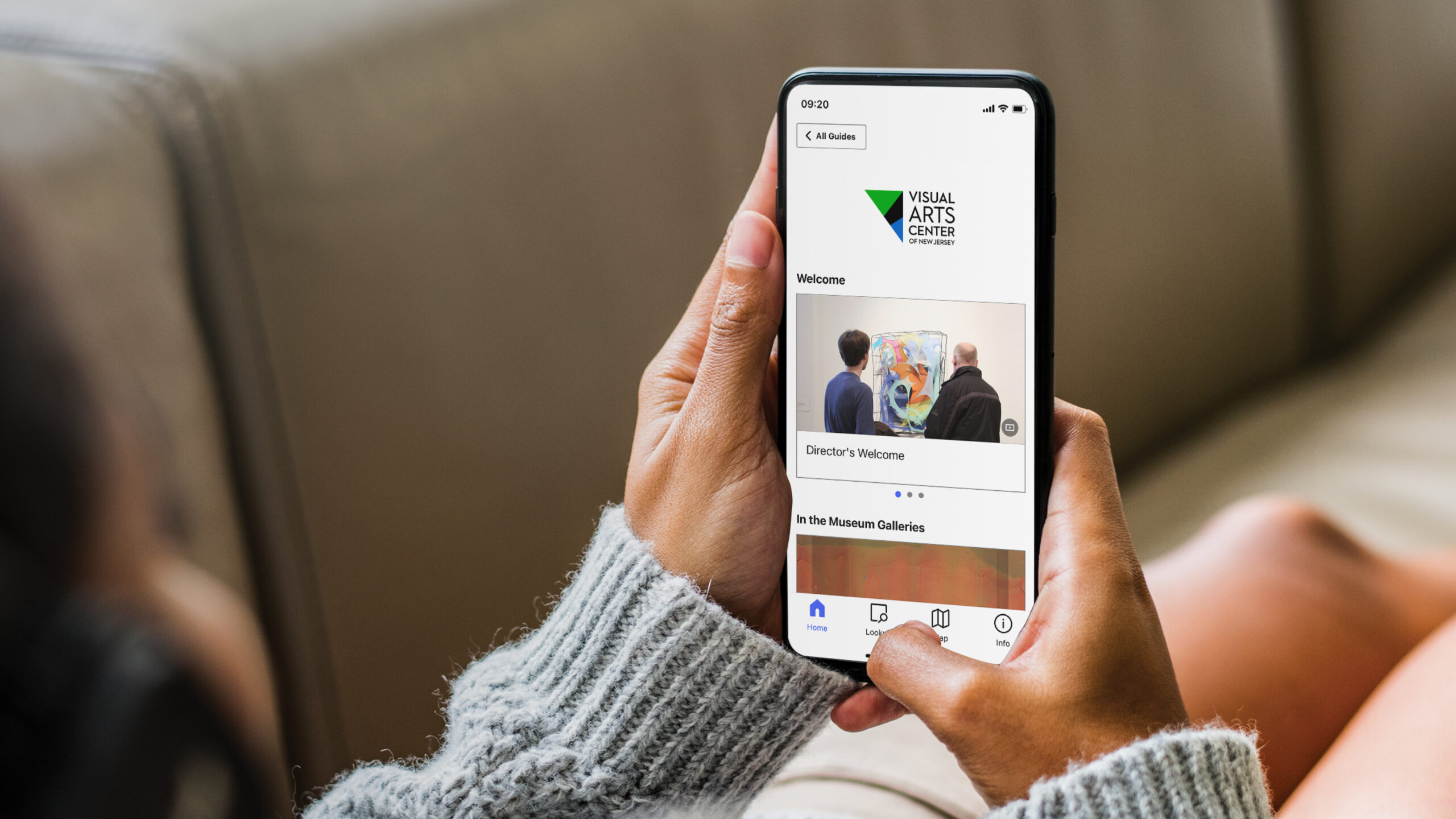 A person wearing a gray sweater holds a smartphone displaying the Visual Arts Center of New Jersey’s mobile guide. The screen shows the Center’s logo and a section titled “Welcome” with a photo of two people viewing a colorful abstract artwork in the gallery.