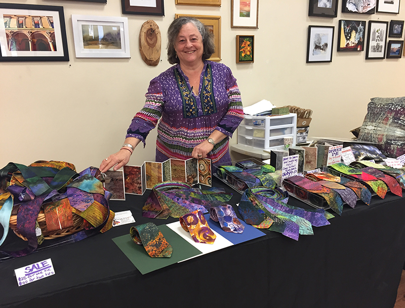 IMG_9687 Feature A smiling woman with gray hair, wearing a purple patterned blouse, stands behind a display table at an art fair. The table is covered with colorful hand-dyed silk ties in a variety of bright patterns and hues, neatly arranged in rows and baskets. Small signs indicate prices and techniques, such as “SALE $25 for one tie, $40 for two ties” and “Disperse Dye.” Behind her, the wall is decorated with framed artwork and photographs.