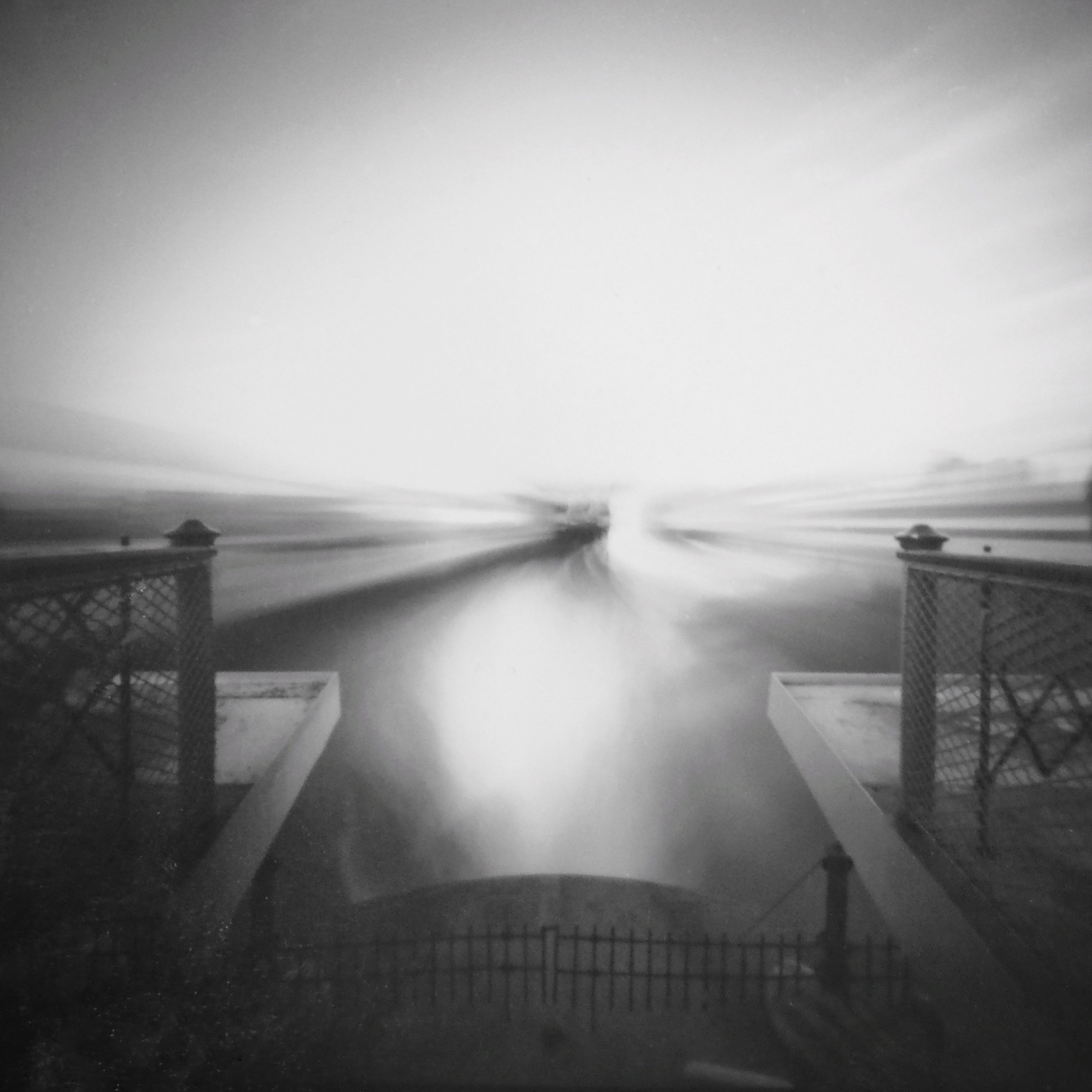 A black-and-white long-exposure photograph taken from a raised platform or bridge, looking out toward a bright, overexposed horizon. Two metal railings with lattice patterns frame the left and right sides, converging toward the center. The light creates a dramatic, streaked effect that blurs the sky and water, giving the scene an ethereal, almost abstract quality. A faint silhouette of a person appears on the right edge, partially blurred due to the long exposure.