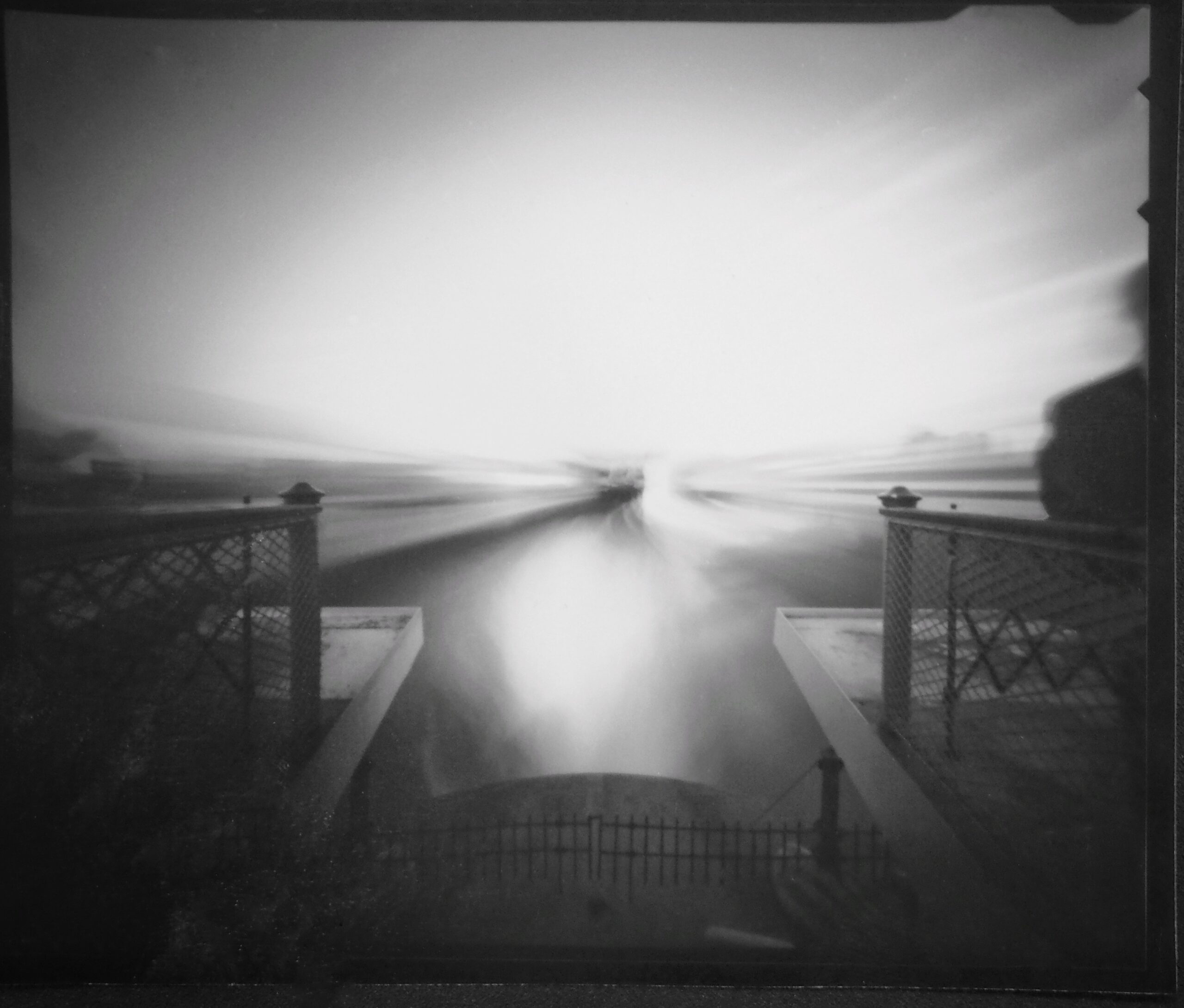 A black-and-white long-exposure photograph taken from a raised platform or bridge, looking out toward a bright, overexposed horizon. Two metal railings with lattice patterns frame the left and right sides, converging toward the center. The light creates a dramatic, streaked effect that blurs the sky and water, giving the scene an ethereal, almost abstract quality. A faint silhouette of a person appears on the right edge, partially blurred due to the long exposure.