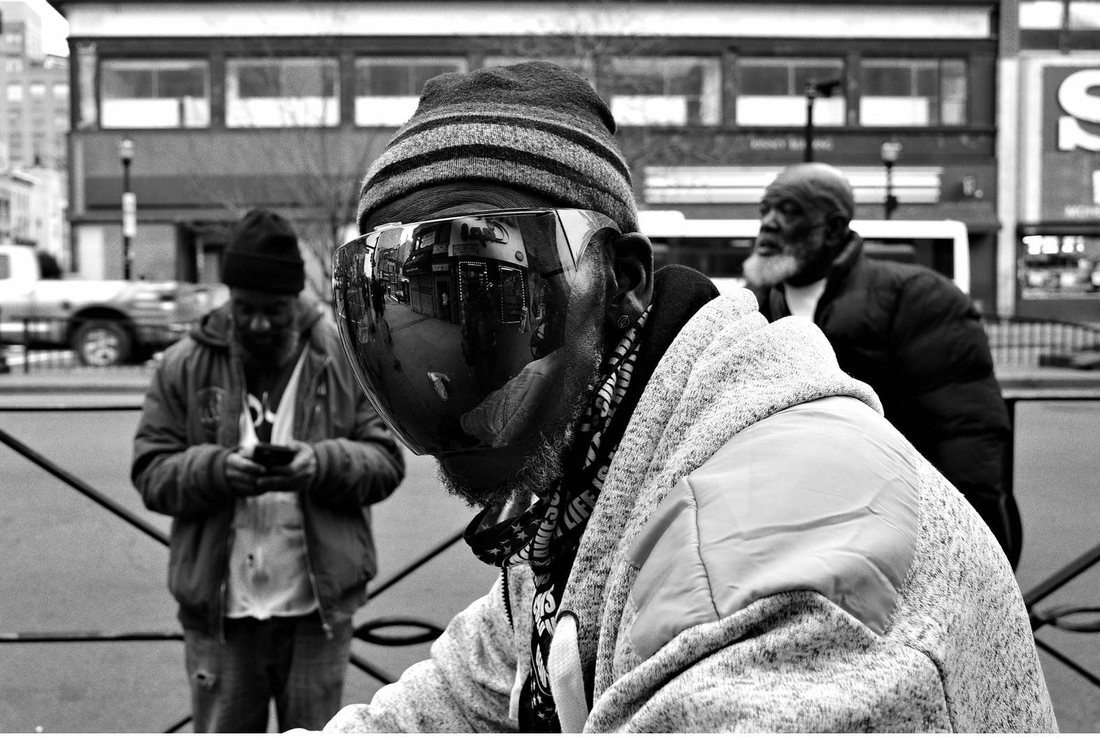Black-and-white street photograph of three men on a city sidewalk. The man in the foreground wears a striped knit hat and an oversized mirrored visor that reflects storefronts and pedestrians. He leans slightly toward the camera. Behind him, one man looks down at his phone and another stands with hands in pockets, glancing away. Urban buildings and cars are out of focus in the background.