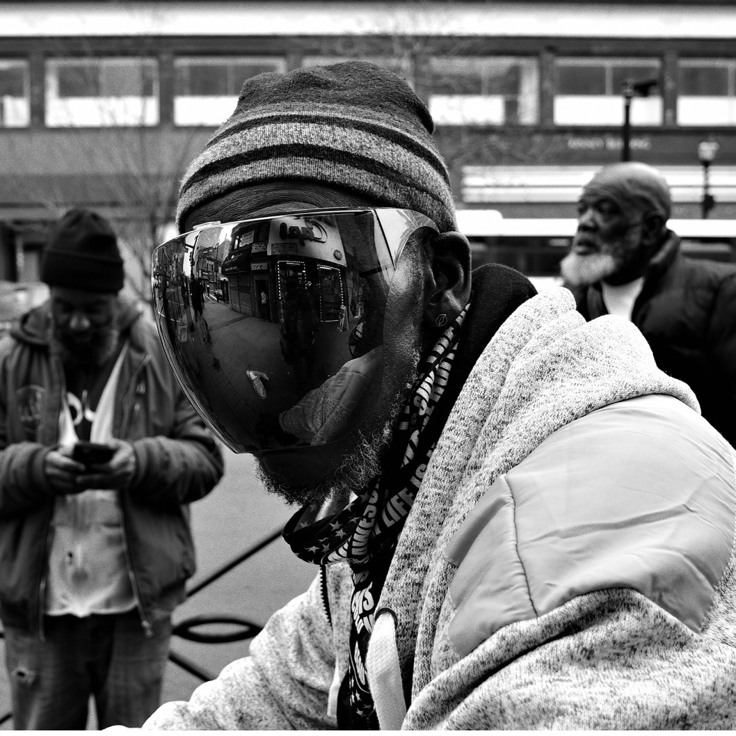 Black-and-white street photograph of three men on a city sidewalk. The man in the foreground wears a striped knit hat and an oversized mirrored visor that reflects storefronts and pedestrians. He leans slightly toward the camera. Behind him, one man looks down at his phone and another stands with hands in pockets, glancing away. Urban buildings and cars are out of focus in the background.