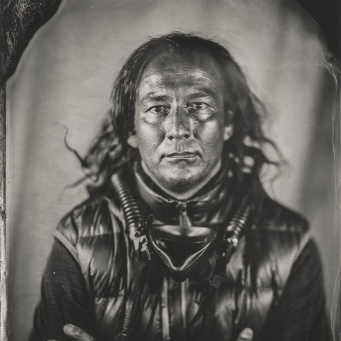 A black-and-white portrait of a man with long hair, looking directly at the camera with a serious expression. He is wearing a shiny, padded vest and has thick cords or breathing tubes draped around his neck and chest. His arms are crossed. The photograph has a wet-plate or tintype style with a soft background and darkened border edges.