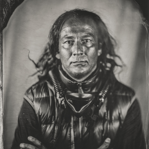 A black-and-white portrait of a man with long hair, looking directly at the camera with a serious expression. He is wearing a shiny, padded vest and has thick cords or breathing tubes draped around his neck and chest. His arms are crossed. The photograph has a wet-plate or tintype style with a soft background and darkened border edges.