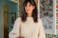 A woman with shoulder-length dark hair and glasses stands indoors holding a clear glass cup. She wears a textured cream-colored sweater and looks slightly off to the side. Behind her are framed abstract artworks on a patterned wall with green leaves and red accents, and an arched doorway leading to another room.