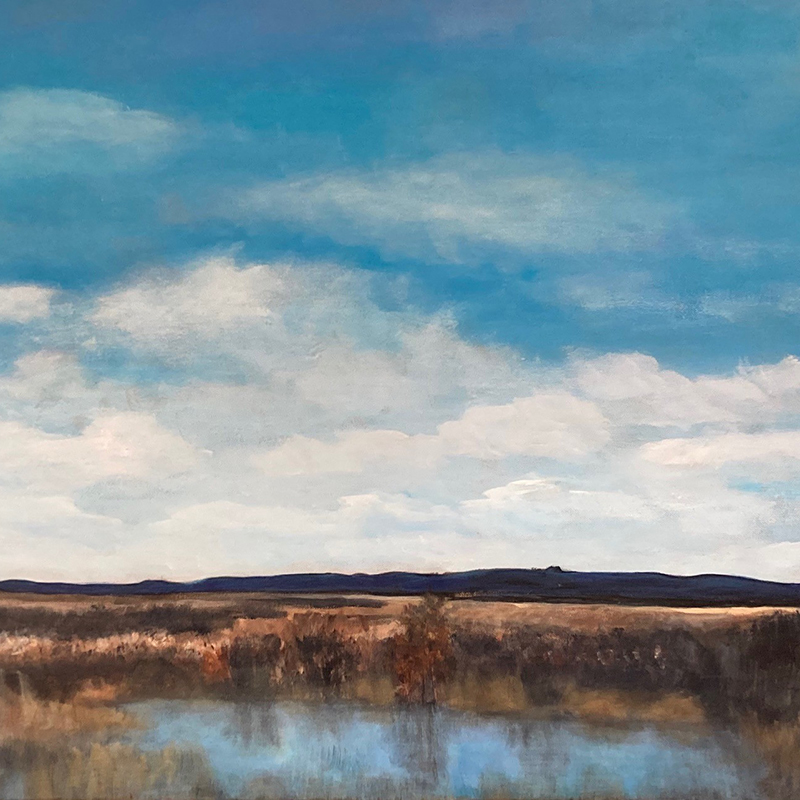A realistic landscape painting depicting a wide, open wetland or marshland scene beneath a dramatic sky. The upper two-thirds of the composition is filled with a deep blue sky and billowing white cumulus clouds. Below, a low ridge of dark hills runs across the horizon. In the foreground, golden-brown marsh grasses and shrubs with touches of rust and amber line the edges of a calm body of water that reflects the blue sky above. The muted earth tones of the wetland contrast with the vibrant sky, creating a serene, expansive mood.