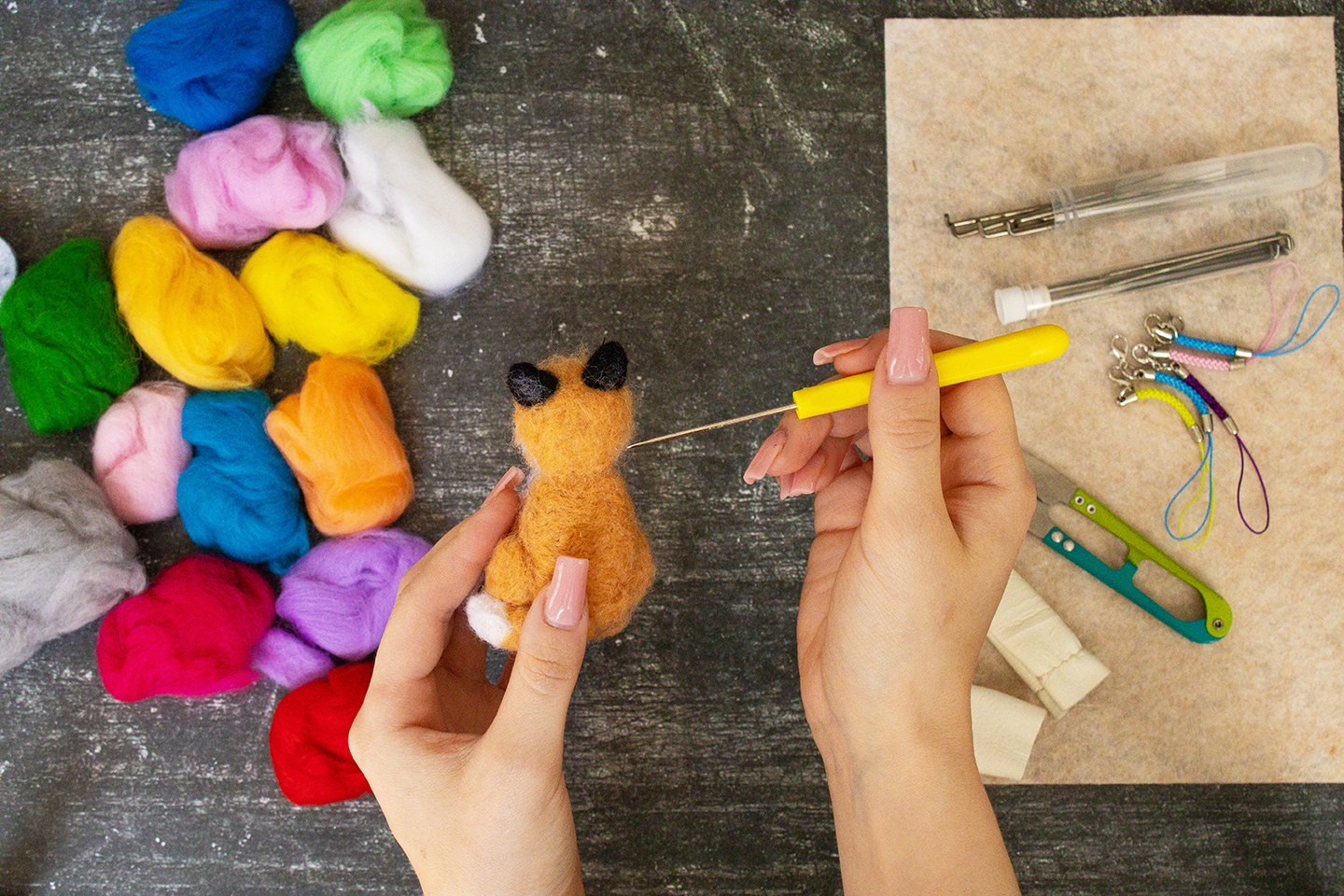 Overhead view of two hands working on a needle-felted fox figurine — one hand holds the small amber-colored fox while the other guides a yellow-handled felting needle into its body. Multicolored balls of wool roving are scattered to the left, and felting tools including needles, clips, and accessories are arranged on a tan foam mat to the right.