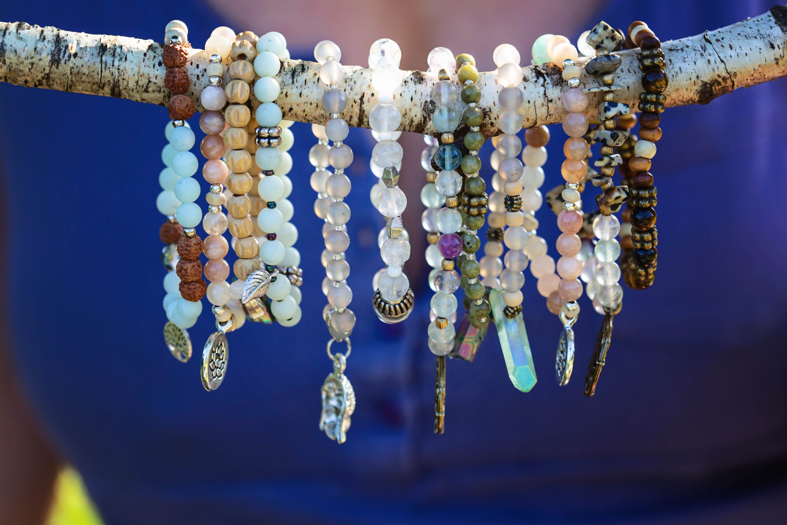 A collection of handmade beaded bracelets displayed on a rustic birch branch against a vibrant cobalt blue background. The bracelets feature an assortment of natural gemstone and wooden beads in soft earth tones — including pale aqua amazonite, peach moonstone, rudraksha seeds, clear quartz, green serpentine, and dalmatian jasper — many adorned with silver and bronze charms such as a tree of life medallion, a leaf, a key, a crystal point, and a feather.