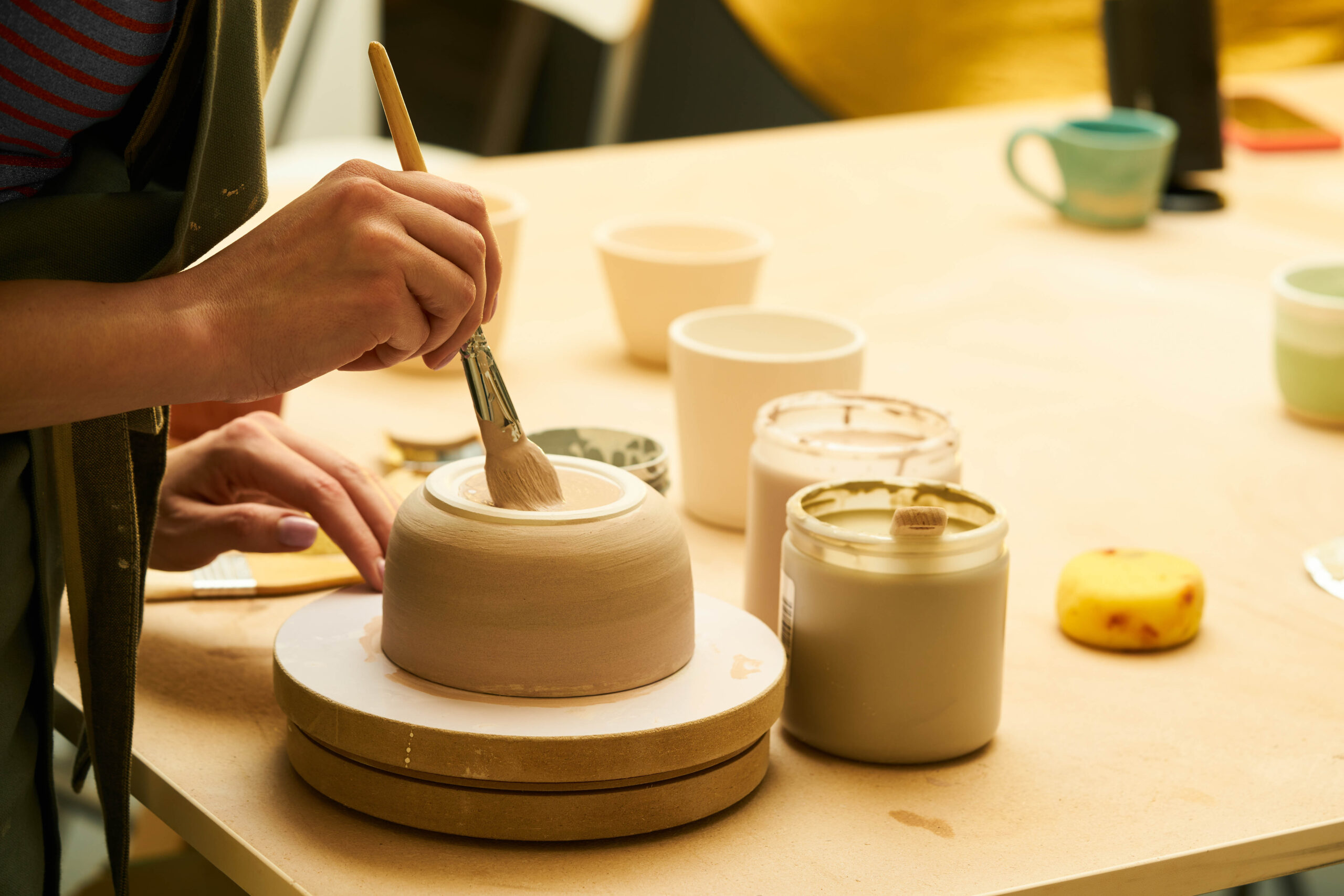 Close-up of girl painting clay mug with glaze. Woman coloring pottery in workshop with a paintbrush. Painter in green apron glazing clay pot.