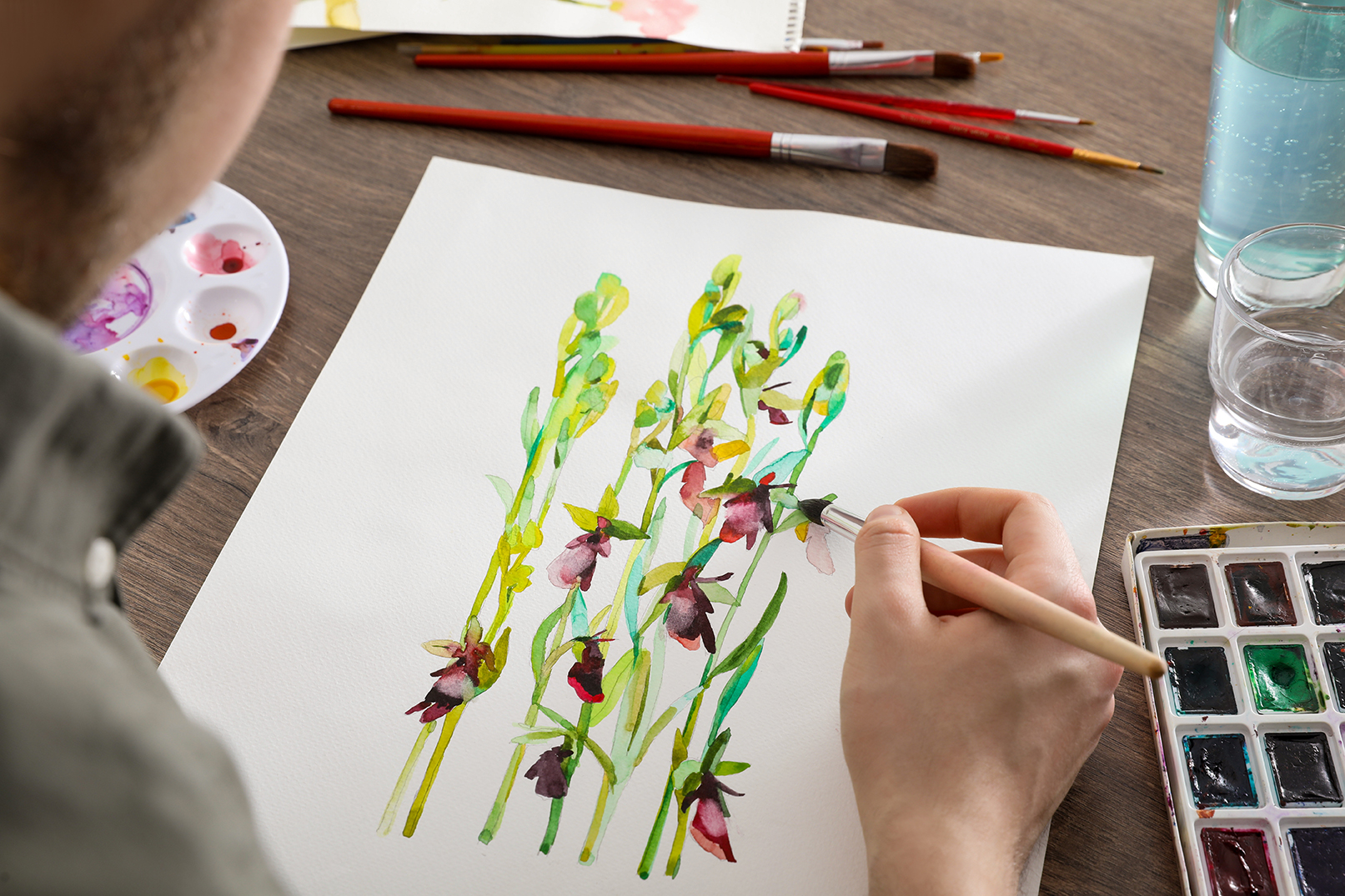 Overhead view of an artist's hand holding a fine brush, adding detail to a watercolor painting of slender flowering stems with dark crimson and green blossoms. Art supplies including red-handled brushes, a mixing palette, and a pan of watercolors are arranged on a wood-grain surface.