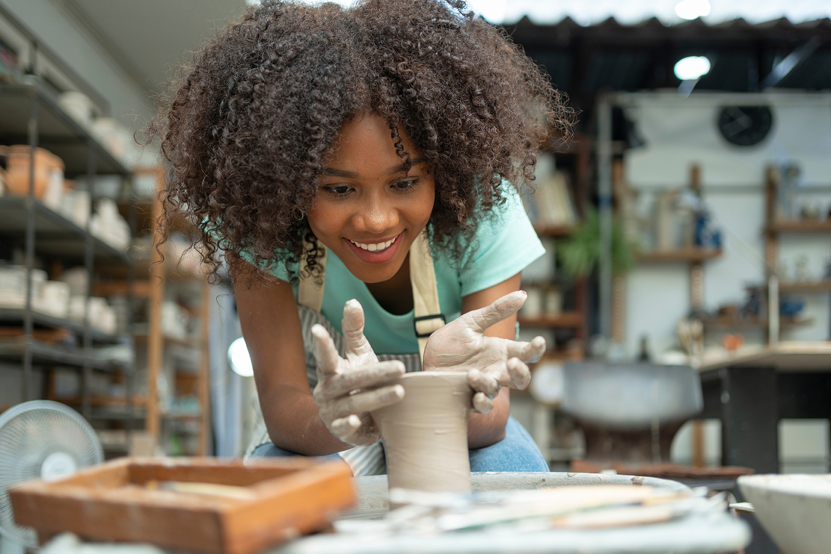 young woman with curly hair leans in with a wide smile as she shapes a clay vessel on a spinning pottery wheel, her hands coated in clay. Shelves of ceramics fill the studio behind her.