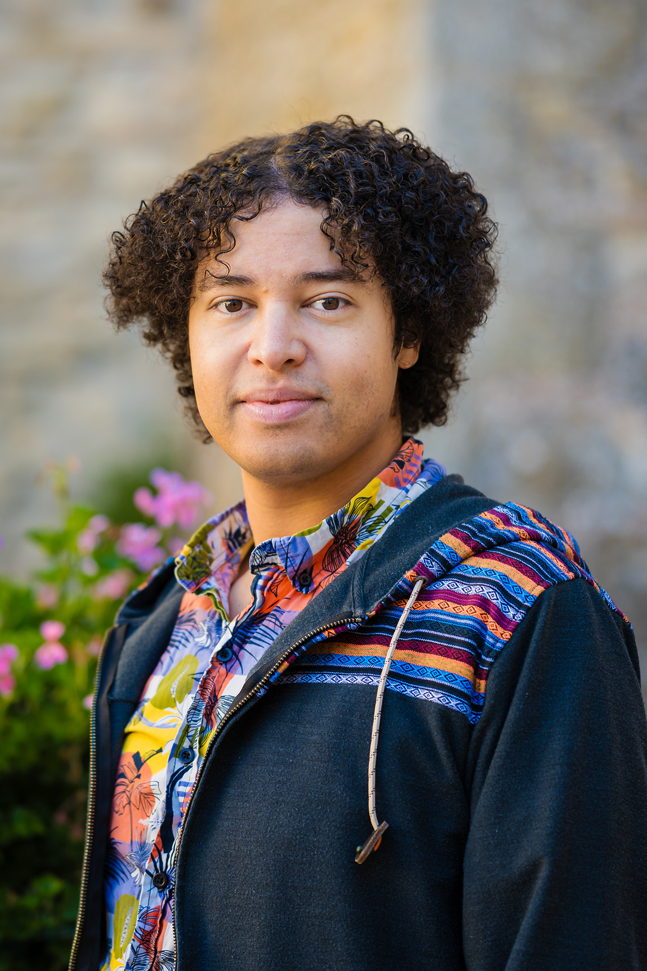 A young person with dark, curly hair stands outdoors in front of a stone wall, with pink and white flowering bushes visible to the left. They are wearing a black zip-up hoodie over a colorful floral shirt and looking calmly at the camera. The photo is a portrait-style shot taken in warm, natural light.