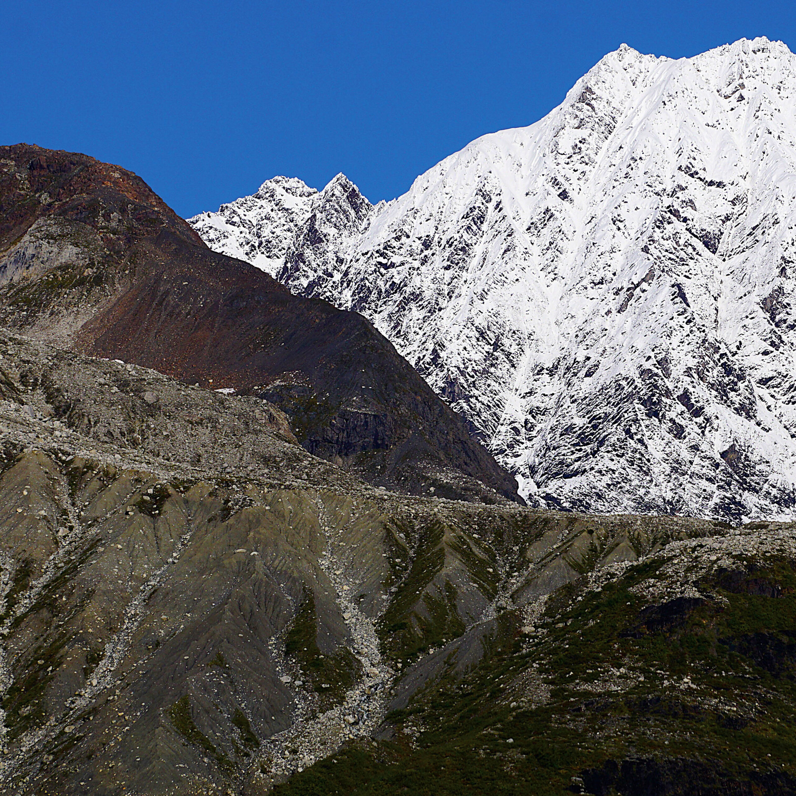 Two mountain peaks rise against a deep blue sky — one a rust-brown rocky summit on the left, the other a massive snow-covered peak dominating the right. In the foreground, eroded grey-green slopes fan out in ridged formations, with sparse alpine vegetation clinging to the lower edges.