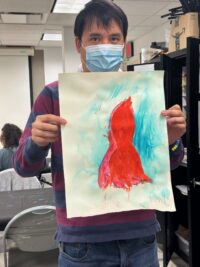 A person wearing a blue surgical mask and a burgundy and navy striped sweater holds up a watercolor painting in what appears to be a classroom or office setting. The painting features an abstract figure — likely a dress or female torso — painted in vivid red and pink against a teal/turquoise brushstroke background on white paper. The artist's signature is visible in the lower right corner of the painting.