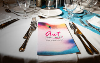 A close-up of an elegantly set round dining table with white linens and teal napkins, featuring a colorful watercolor program booklet reading 'art EMPOWERS! 2026 Spring Gala' from the Visual Arts Center of New Jersey, surrounded by silver cutlery and crystal glassware.