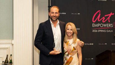 Two people pose together in front of a step-and-repeat backdrop for the 'art EMPOWERS! 2026 Spring Gala' presented by the Visual Arts Center of New Jersey, with sponsors Chubb and Brown & Brown Risk visible. The man on the left holds a glass award trophy.