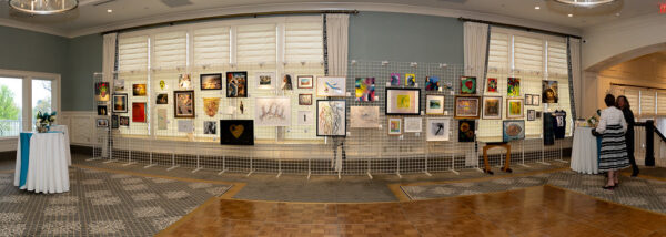 A panoramic view of a silent auction art exhibition inside an elegant event venue, with dozens of framed artworks displayed on white wire grid panels along a windowed wall. Two attendees browse the artwork on the right side of the room, and a teal-accented cocktail table is visible on the left.