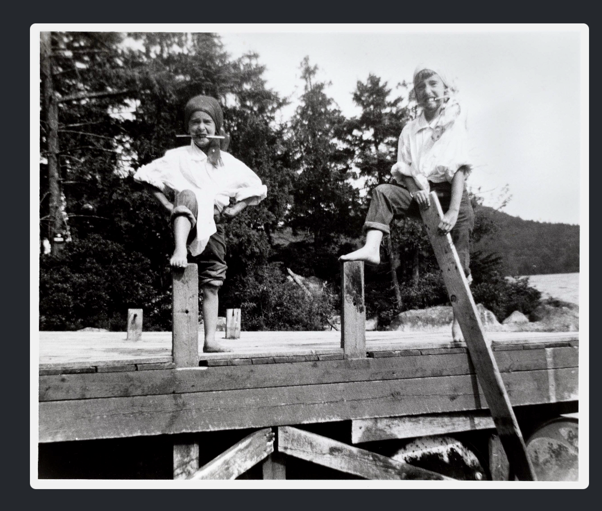 A vintage black-and-white photograph — two children (titled "Geoffrey and his sister as pirates") sitting on wooden posts on a dock or pier, dressed in playful pirate-style costumes with headscarves, one biting a "knife" prop, near a lake with trees in the background.