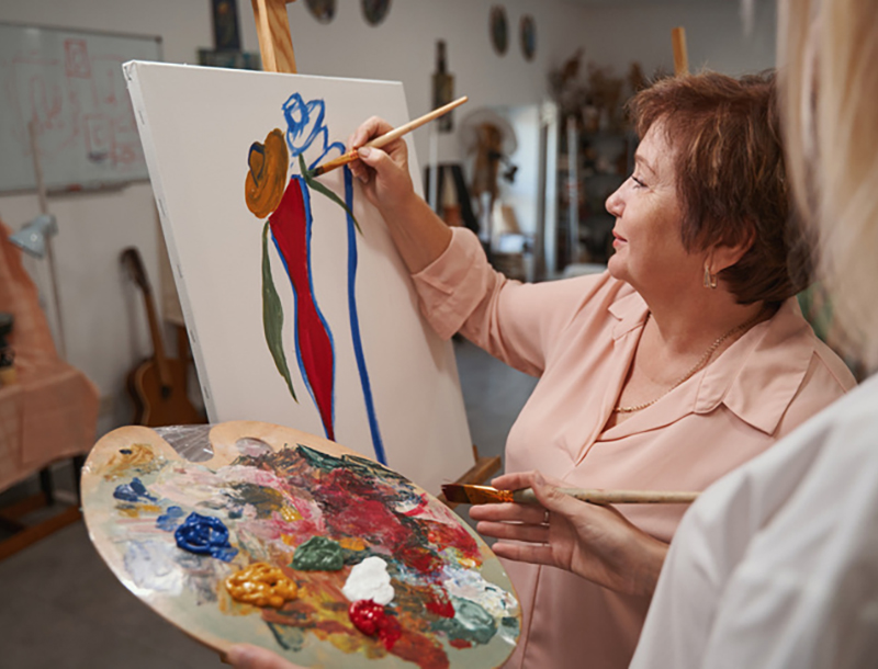 An older woman with short auburn hair, wearing a light pink blouse, smiles as she paints a colorful abstract flower on a canvas at an easel. She holds a paint-covered palette and a second brush, while another person looks on in the foreground of an art studio.